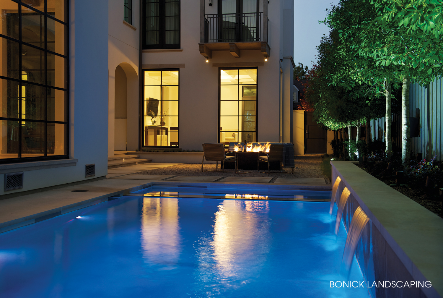 Night shot of pool in foreground, fire pit in background.